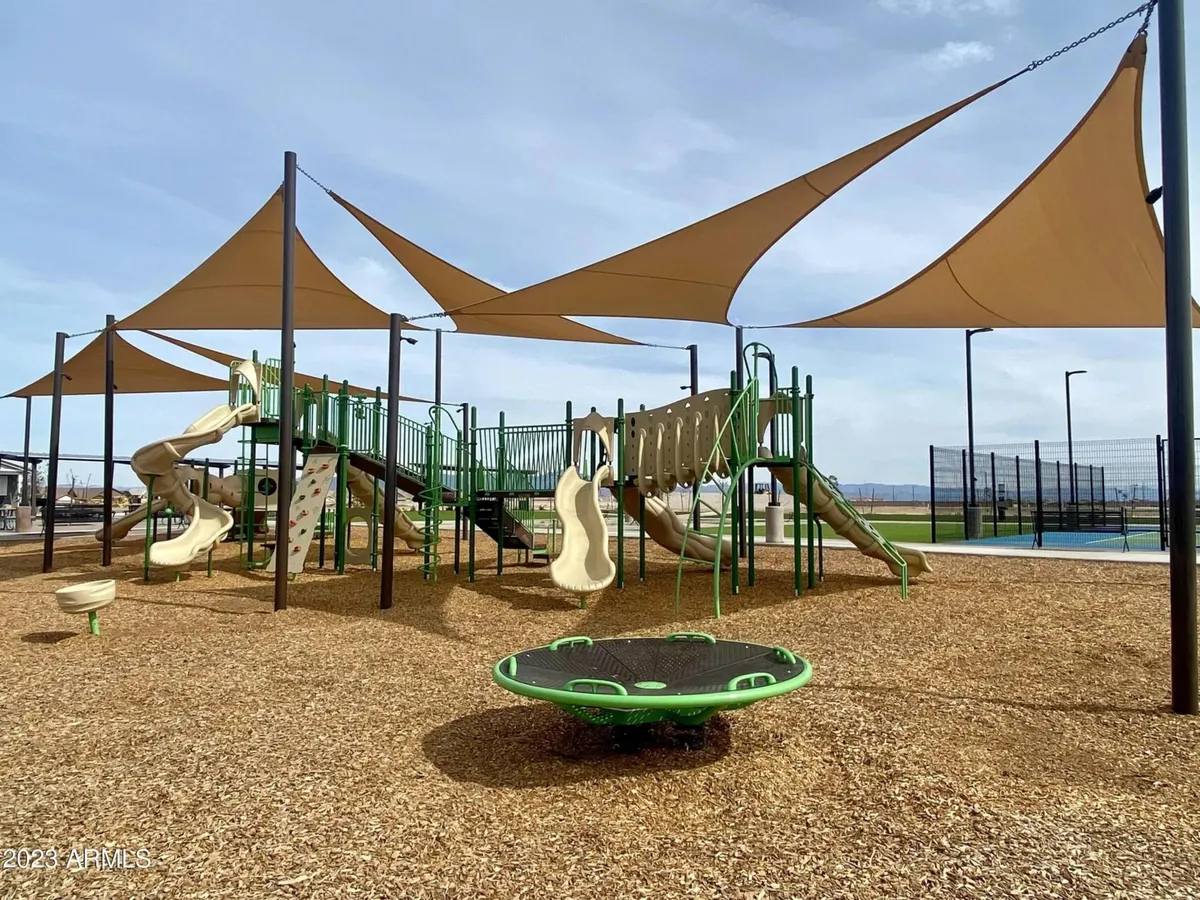 The scene features a colorful playground with shade sails, sand area, and a sport court in the background. Families and children can be seen enjoying outdoor play, while the surrounding area suggests a welcoming community setting.