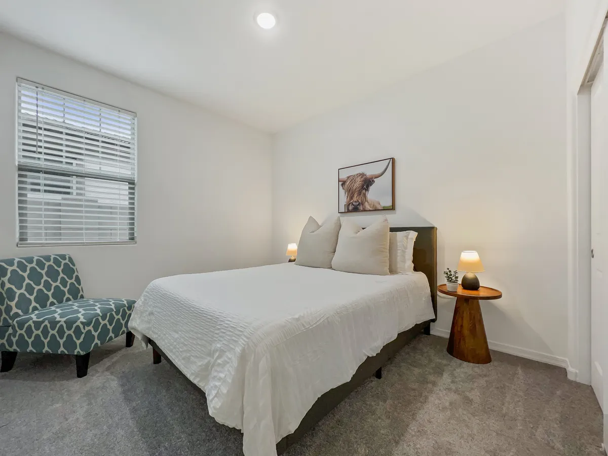 A serene bedroom featuring a bed with white linens, decorative pillows, soft artwork on the wall, an accent chair, and a nightstand with lamps. Natural light gently fills the space, creating a peaceful atmosphere ideal for rest and relaxation.