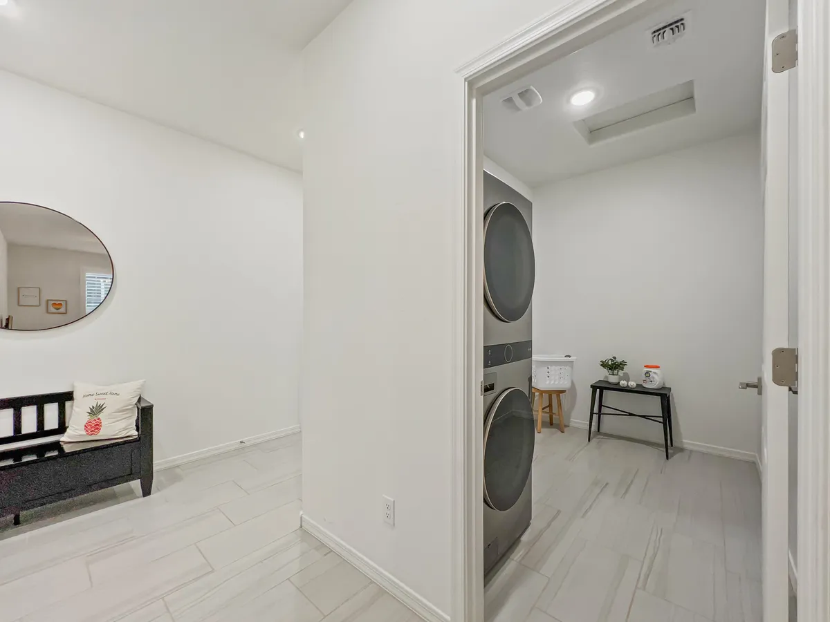 A bright and functional laundry room featuring modern laundry machines, an oval mirror, a black bench for seating or folding, and a small table with plants for a welcoming touch. The space feels organized and comfortable, making chores more pleasant.
