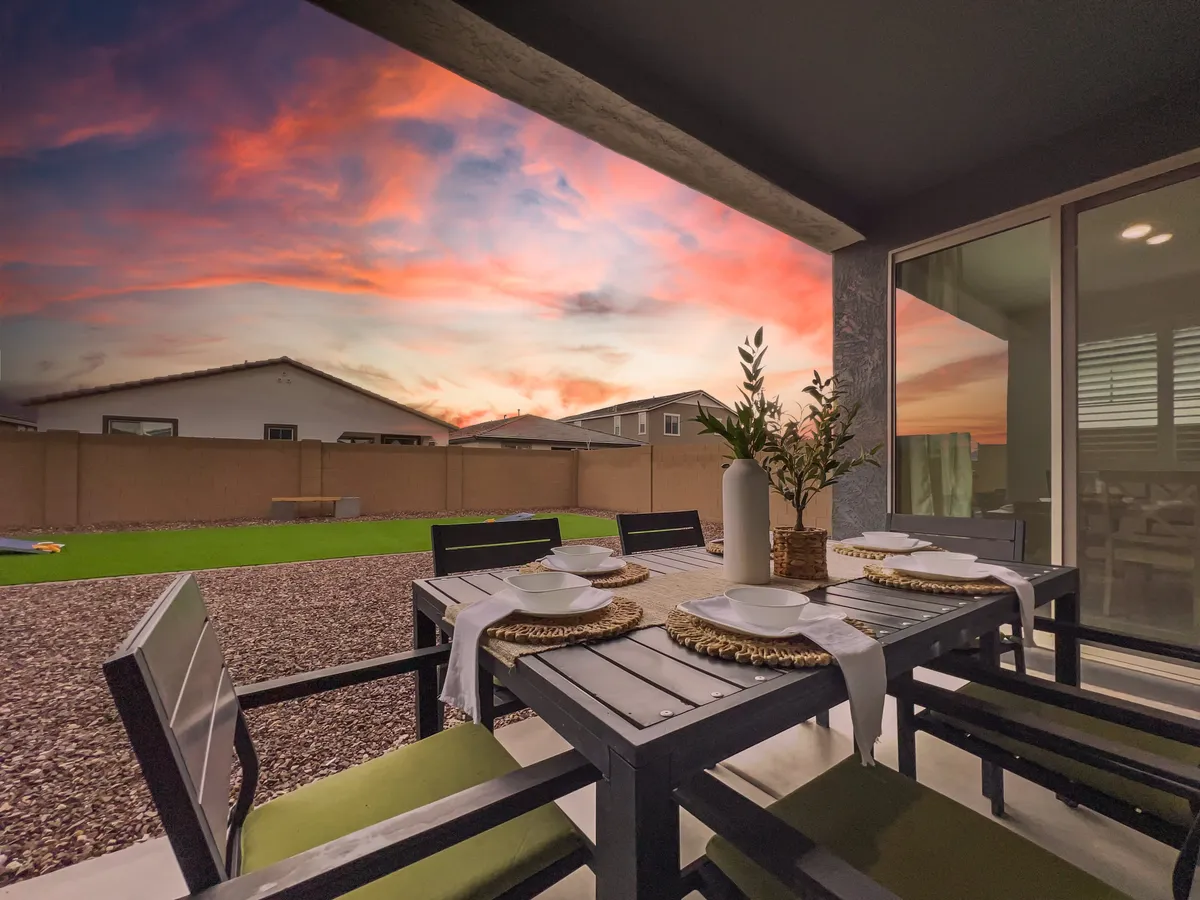 A serene backyard scene with a dining table set for a meal, plates and cutlery arranged, a vase with greenery, and the colorful sunset sky in the background. The space suggests relaxation, dining, and social gatherings in a private outdoor setting.