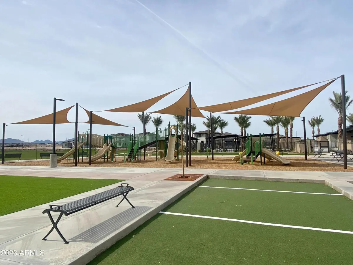 A vibrant neighborhood playground scene with colorful equipment, shade sails overhead, benches for families, and palm trees in the background. Children are actively playing, and the atmosphere is lively, hinting at joyful community interactions and family-friendly fun.