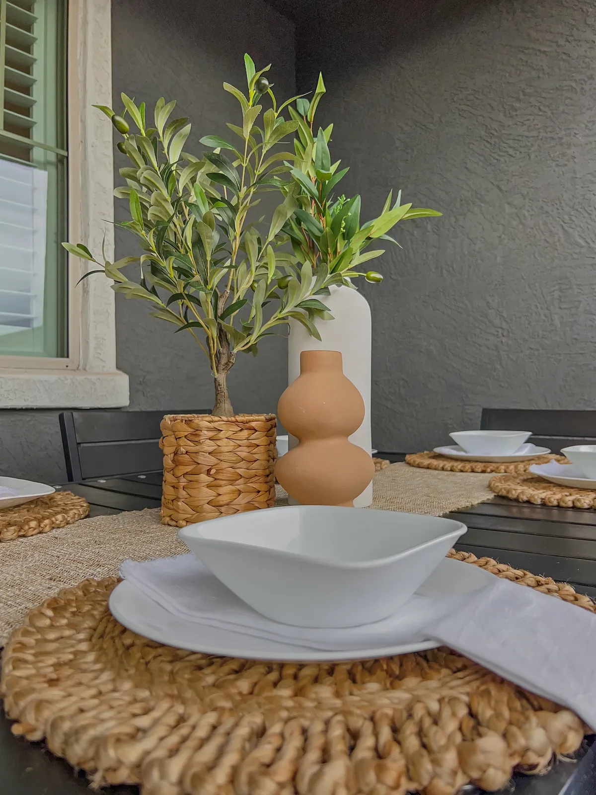 This image showcases an inviting outdoor dining setup with greenery, decorative vases, woven placemats, and white dishware. The scene captures the atmosphere of al fresco gatherings—ideal for barbecues, celebrations, or shared meals under the sun.