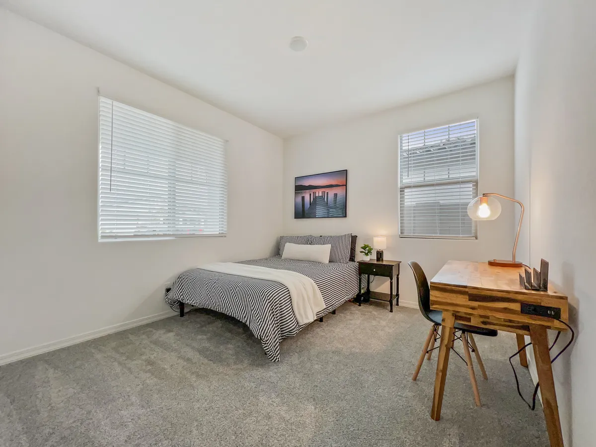 A tranquil bedroom with striped bedding, a wooden desk and lamp set up as a workspace, and calming wall art. The environment feels tailored for both productivity and relaxation, with a cozy reading nook by the window.