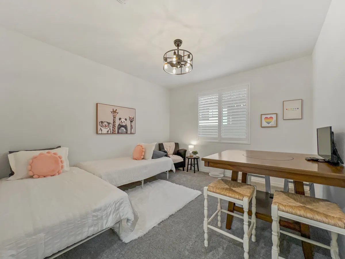 A cheerful interior scene featuring two twin beds, a wooden table, artwork on the wall, and shuttered windows. The space feels bright, welcoming, and clearly designed to accommodate both play and relaxation for children or families. The setup encourages a variety of activities, such as games or creative projects, and is visually inviting for all ages.