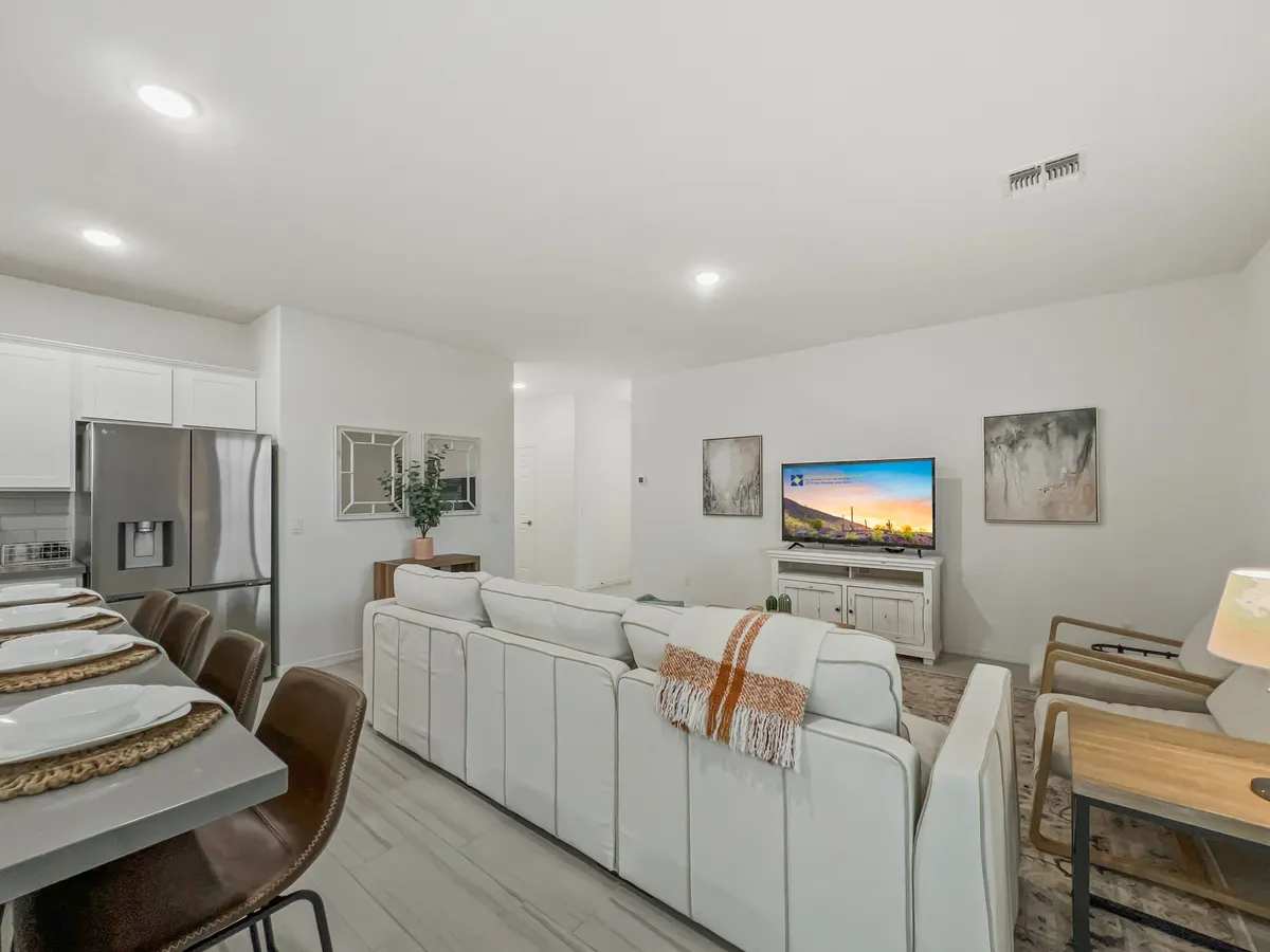 This image captures a spacious open-plan living area featuring a sectional sofa, coffee table, oversized TV, dining area, kitchen appliances, and decorative plants. The layout fosters a communal feeling, with seamless transitions between spaces for relaxing, dining, and entertaining.